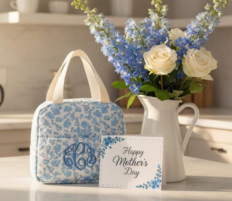 Może przedstawiać: Torba na lunch z kwiatowym nadrukiem i monogramem, bukiet niebieskich ostr&oacute;żek i białych r&oacute;ż w białym dzbanku oraz kartka z napisem "Happy Mother's Day". Torba na lunch ma jasnoniebieski kwiatowy wz&oacute;r.