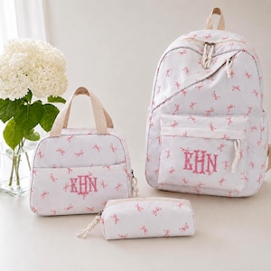 Pode incluir: Um conjunto de três malas a condizer: uma mochila, uma lancheira e um estojo. Todas são brancas com um padrão de laço rosa e um monograma rosa. A mochila tem uma pega superior e um bolso frontal.