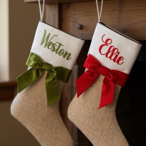Peut inclure: Deux chaussettes de Noël en toile de jute beige avec des revers blancs. La chaussette de gauche porte le nom Weston brodé en vert avec un nœud vert. La chaussette de droite porte le nom Ellie brodé en rouge avec un nœud rouge. Elles sont suspendues par des boucles blanches.
