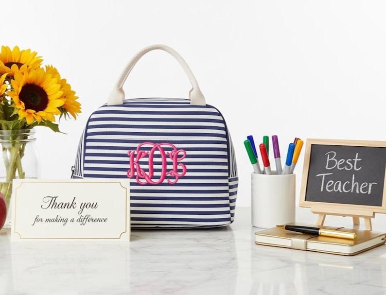 Może przedstawiać: Granatowo-biała torba na lunch w paski z r&oacute;żowym monogramem. Mała tablica z napisem "Best Teacher" stoi obok notatnika i długopis&oacute;w. Obecny jest r&oacute;wnież bukiet słonecznik&oacute;w i kartka z podziękowaniami.