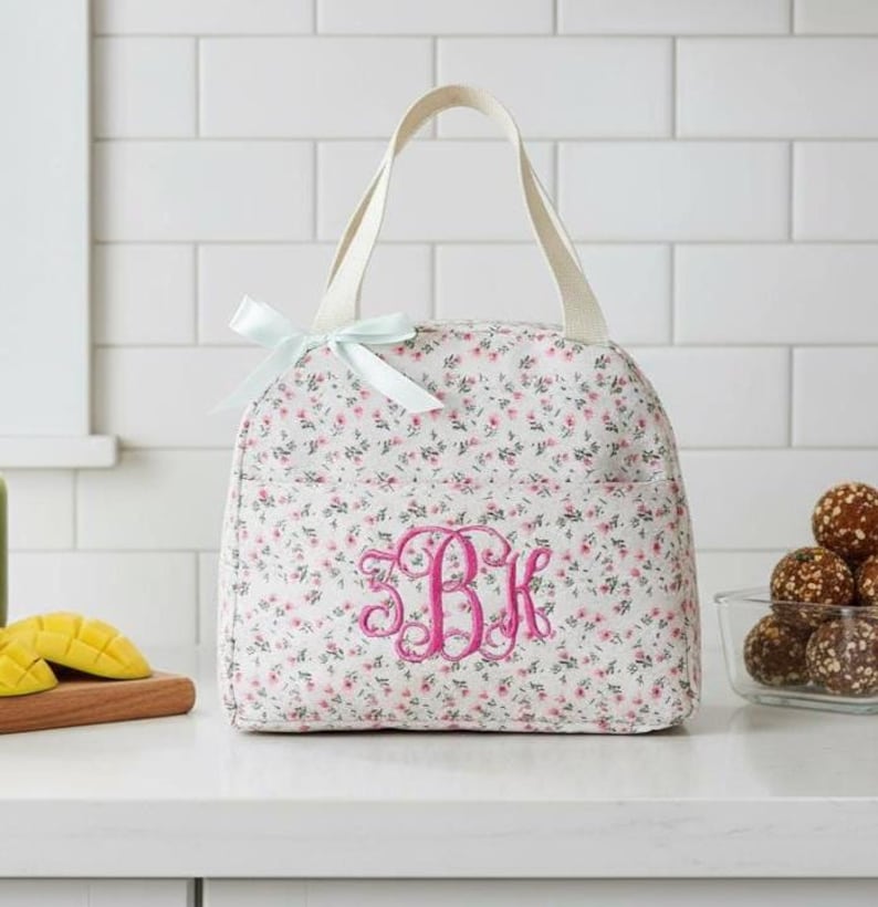 Może przedstawiać: Biała torba na lunch z kwiatowym nadrukiem, jasnoniebieską kokardą i r&oacute;żowym monogramem. Torba ma uchwyt i kieszeń z boku. Torba stoi na białym blacie.