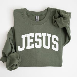 Puede incluir: Sudadera verde oliva con la palabra "JESUS" en letras blancas y gruesas en el pecho. Las mangas largas est&aacute;n dobladas. La sudadera est&aacute; hecha de un material suave.