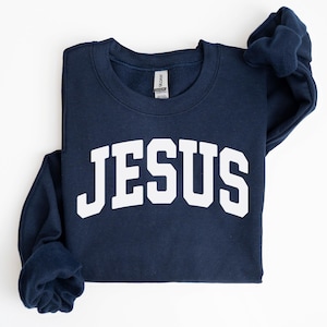 Puede incluir: Sudadera azul marino con la palabra "JESUS" en letras blancas grandes en el pecho. La sudadera est&aacute; doblada cuidadosamente sobre una superficie blanca. Las mangas est&aacute;n dobladas.