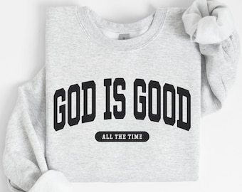 May include: A pale grey sweatshirt featuring the words "GOD IS GOOD" in large, black, curved lettering. Beneath, in a smaller black rectangle, is the phrase "ALL THE TIME". The sweatshirt is folded neatly.