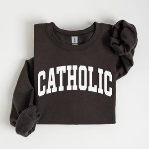 Peut inclure: Un sweat-shirt gris fonc&eacute; avec le mot "CATHOLIC" imprim&eacute; en grandes lettres blanches arqu&eacute;es sur la poitrine. Les manches longues sont pli&eacute;es. Le sweat-shirt est fait d'une mati&egrave;re douce.