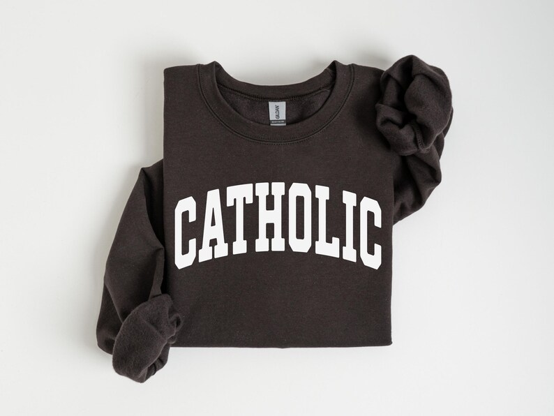 Peut inclure: Un sweat-shirt gris fonc&eacute; avec le mot "CATHOLIC" imprim&eacute; en grandes lettres blanches arqu&eacute;es sur la poitrine. Les manches longues sont pli&eacute;es. Le sweat-shirt est fait d'une mati&egrave;re douce.