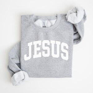 Puede incluir: Sudadera gris jaspeado con la palabra "JESUS" impresa en letras blancas grandes en la parte delantera. Las mangas est&aacute;n dobladas, revelando el interior blanco. Prenda casual y c&oacute;moda.