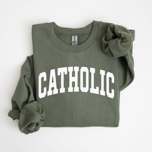 Peut inclure: Sweat-shirt vert olive avec le mot "CATHOLIC" imprim&eacute; en blanc sur la poitrine. Les manches longues sont pli&eacute;es. Le sweat-shirt est en mati&egrave;re douce.