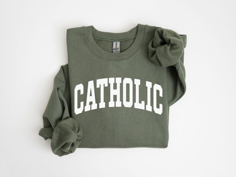 Peut inclure: Sweat-shirt vert olive avec le mot "CATHOLIC" imprim&eacute; en blanc sur la poitrine. Les manches longues sont pli&eacute;es. Le sweat-shirt est en mati&egrave;re douce.