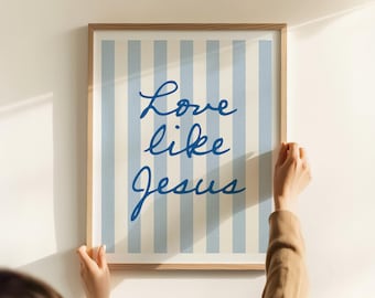 May include: A framed print with a light wood frame. The artwork features a striped background in light blue and white, with the words "Love like Jesus" in blue cursive script. The print is being held by two hands.