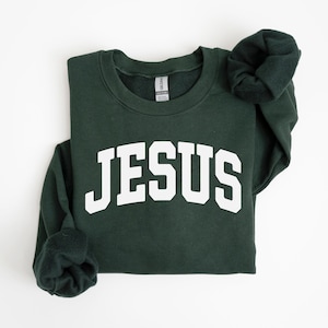 Puede incluir: Una sudadera verde oscuro con la palabra "JESUS" en letras blancas grandes en el pecho. La sudadera tiene cuello redondo y mangas largas, con las mangas dobladas. El fondo es blanco.