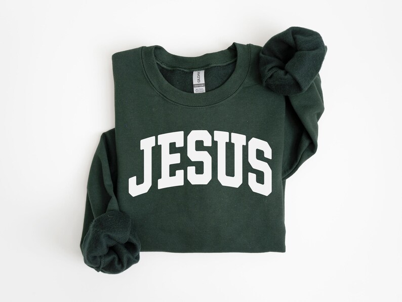 Puede incluir: Una sudadera verde oscuro con la palabra "JESUS" en letras blancas grandes en el pecho. La sudadera tiene cuello redondo y mangas largas, con las mangas dobladas. El fondo es blanco.