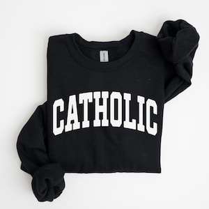 Peut inclure: Sweat-shirt noir avec le mot "CATHOLIC" imprim&eacute; en grandes lettres blanches sur la poitrine. Le sweat-shirt est pli&eacute;, avec les manches rentr&eacute;es. Le fond est blanc uni.