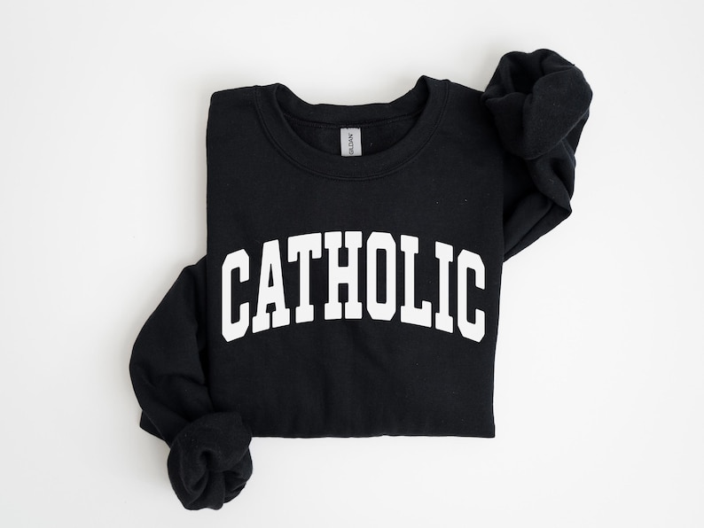 Peut inclure: Sweat-shirt noir avec le mot "CATHOLIC" imprim&eacute; en grandes lettres blanches sur la poitrine. Le sweat-shirt est pli&eacute;, avec les manches rentr&eacute;es. Le fond est blanc uni.