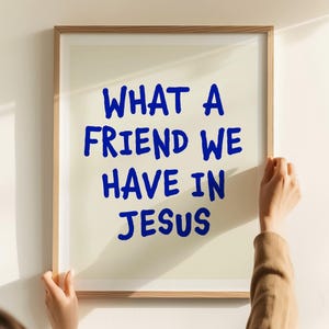 May include: A framed print with the text "WHAT A FRIEND WE HAVE IN JESUS" in blue lettering. The frame is a light brown color. The print is being held by a person.