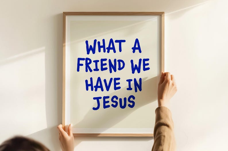 May include: A framed print with the text "WHAT A FRIEND WE HAVE IN JESUS" in blue lettering. The frame is a light brown color. The print is being held by a person.