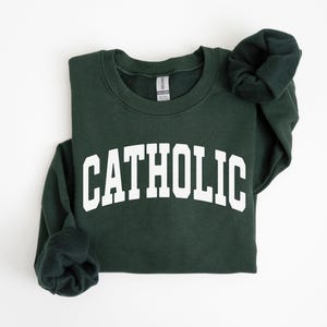 Peut inclure: Un sweat-shirt vert fonc&eacute; avec le mot "CATHOLIC" imprim&eacute; en lettres blanches et grasses. Le sweat-shirt est pli&eacute;, avec les manches repli&eacute;es. Le fond est blanc uni.