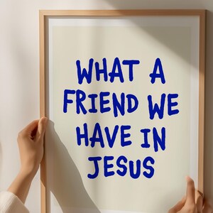 May include: A framed print with the text "WHAT A FRIEND WE HAVE IN JESUS" in blue, handwritten-style font. The print is held by a person, with a neutral background and a wooden frame.