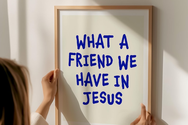 May include: A framed print with the text "WHAT A FRIEND WE HAVE IN JESUS" in blue, handwritten-style font. The print is held by a person, with a neutral background and a wooden frame.