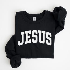 Puede incluir: Sudadera negra de cuello redondo con la palabra "JES&Uacute;S" en letras grandes y blancas en el pecho. La sudadera est&aacute; doblada, con las mangas cuidadosamente dispuestas. El fondo es blanco liso.