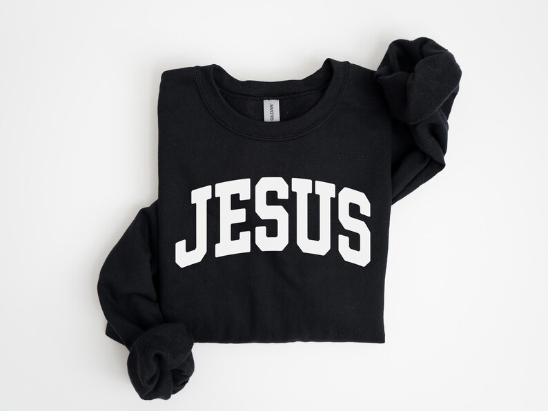 Puede incluir: Sudadera negra de cuello redondo con la palabra "JES&Uacute;S" en letras grandes y blancas en el pecho. La sudadera est&aacute; doblada, con las mangas cuidadosamente dispuestas. El fondo es blanco liso.