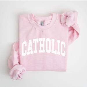 Peut inclure: Un sweat-shirt rose clair avec le mot "CATHOLIC" imprim&eacute; en grandes lettres blanches sur la poitrine. Le sweat-shirt a un col rond et des manches longues, avec les manches repli&eacute;es. Le fond est blanc.