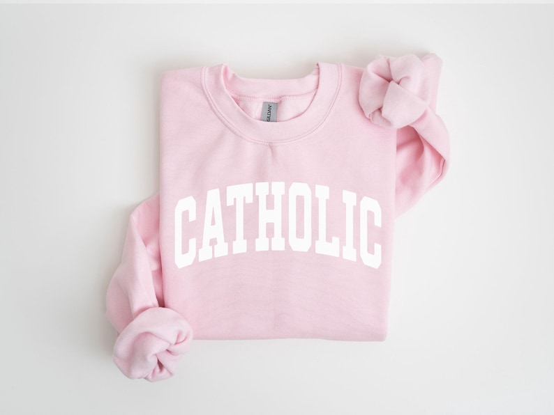 Peut inclure: Un sweat-shirt rose clair avec le mot "CATHOLIC" imprim&eacute; en grandes lettres blanches sur la poitrine. Le sweat-shirt a un col rond et des manches longues, avec les manches repli&eacute;es. Le fond est blanc.