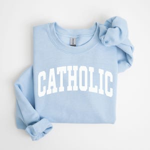 Peut inclure: Un sweat-shirt bleu clair avec le mot "CATHOLIC" imprim&eacute; en grandes lettres blanches. Le sweat-shirt est pli&eacute; proprement, avec les manches et la capuche visibles. Le fond est blanc uni.
