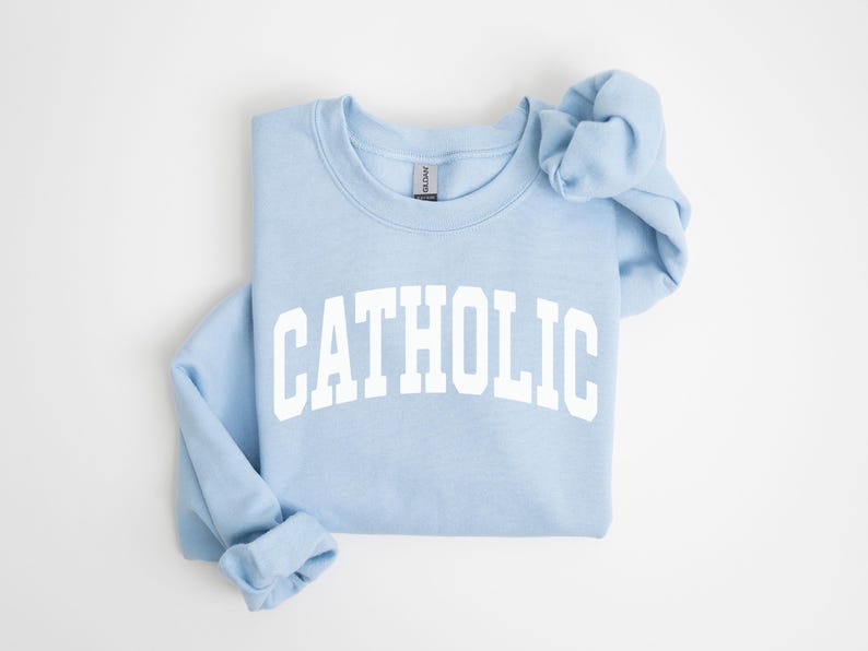 Peut inclure: Un sweat-shirt bleu clair avec le mot "CATHOLIC" imprim&eacute; en grandes lettres blanches. Le sweat-shirt est pli&eacute; proprement, avec les manches et la capuche visibles. Le fond est blanc uni.
