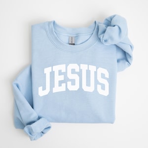 Puede incluir: Sudadera azul claro con la palabra "JESUS" en letras blancas grandes en el pecho. La sudadera est&aacute; doblada cuidadosamente, con las mangas y el cuello visibles. La prenda es de un material suave.