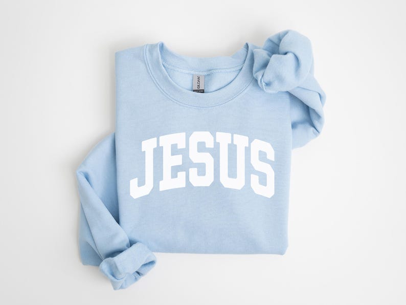 Puede incluir: Sudadera azul claro con la palabra "JESUS" en letras blancas grandes en el pecho. La sudadera est&aacute; doblada cuidadosamente, con las mangas y el cuello visibles. La prenda es de un material suave.