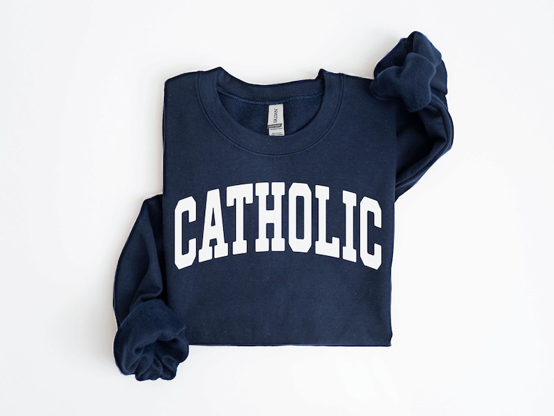 Peut inclure: Sweat-shirt bleu marine avec le mot "CATHOLIC" imprim&eacute; en grandes lettres blanches sur la poitrine. Le sweat-shirt a un col rond et des manches longues. Le v&ecirc;tement est pli&eacute; sur une surface blanche.