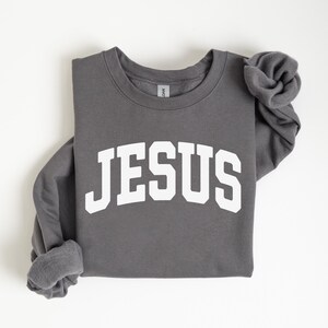 Puede incluir: Una sudadera gris oscuro con la palabra "JESUS" impresa en letras blancas grandes en la parte delantera. Las mangas est&aacute;n dobladas. La sudadera est&aacute; hecha de un material suave.