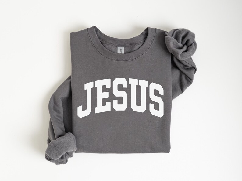 Puede incluir: Una sudadera gris oscuro con la palabra "JESUS" impresa en letras blancas grandes en la parte delantera. Las mangas est&aacute;n dobladas. La sudadera est&aacute; hecha de un material suave.