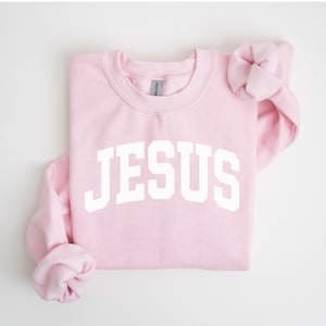 Puede incluir: Una sudadera rosa claro con la palabra "JESUS" impresa en letras blancas en el pecho. Las mangas est&aacute;n dobladas y la sudadera est&aacute; colocada sobre una superficie blanca. La sudadera es de cuello redondo.