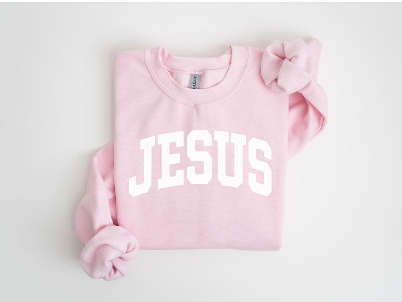 Puede incluir: Una sudadera rosa claro con la palabra "JESUS" impresa en letras blancas en el pecho. Las mangas est&aacute;n dobladas y la sudadera est&aacute; colocada sobre una superficie blanca. La sudadera es de cuello redondo.