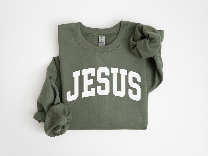 Puede incluir: Sudadera verde oliva con la palabra "JESUS" en letras blancas y gruesas en el pecho. Las mangas largas est&aacute;n dobladas. La sudadera est&aacute; hecha de un material suave.