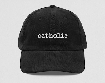 May include: Black corduroy baseball cap with the word "catholic" embroidered in white across the front. The cap has a curved brim and an adjustable strap at the back.