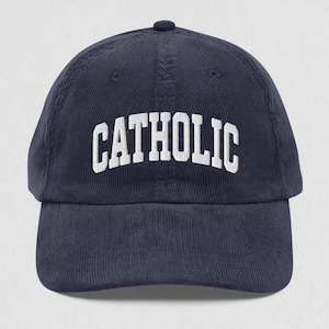 Op de afbeelding: Marineblauwe corduroy baseballcap met het woord "CATHOLIC" in witte blokletters geborduurd op de voorkant. De cap heeft een gebogen klep en een kleine knoop bovenop.