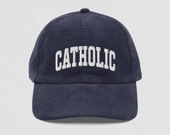 May include: Navy blue corduroy baseball cap with the word "CATHOLIC" embroidered in white block letters across the front. The cap has a curved brim and a small button on top.