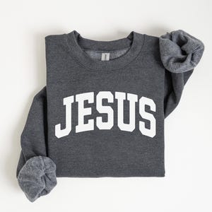 Puede incluir: Una sudadera gris oscuro con la palabra "JESUS" impresa en letras blancas grandes y arqueadas. Las mangas largas est&aacute;n dobladas. La sudadera est&aacute; doblada cuidadosamente sobre una superficie blanca.