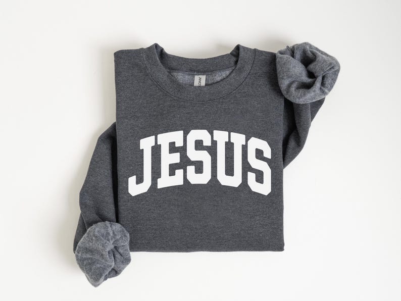 Puede incluir: Una sudadera gris oscuro con la palabra "JESUS" impresa en letras blancas grandes y arqueadas. Las mangas largas est&aacute;n dobladas. La sudadera est&aacute; doblada cuidadosamente sobre una superficie blanca.