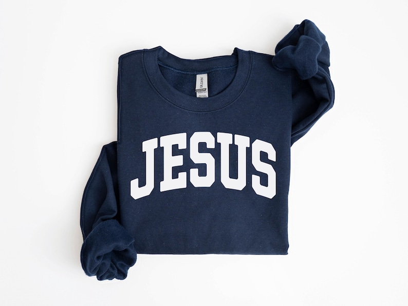 Puede incluir: Sudadera azul marino con la palabra "JESUS" en letras blancas grandes en el pecho. La sudadera est&aacute; doblada cuidadosamente sobre una superficie blanca. Las mangas est&aacute;n dobladas.
