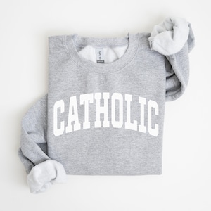 Peut inclure: Un sweat-shirt gris chin&eacute; avec le mot "CATHOLIC" imprim&eacute; en grandes lettres blanches arqu&eacute;es. Le sweat-shirt &agrave; col rond a des manches longues, avec les poignets repli&eacute;s pour r&eacute;v&eacute;ler une doublure gris clair.