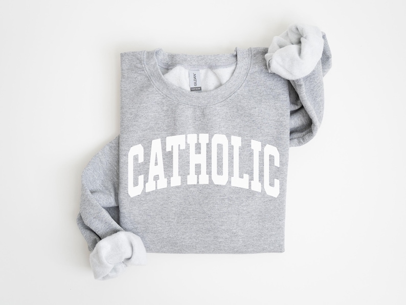 Peut inclure: Un sweat-shirt gris chin&eacute; avec le mot "CATHOLIC" imprim&eacute; en grandes lettres blanches arqu&eacute;es. Le sweat-shirt &agrave; col rond a des manches longues, avec les poignets repli&eacute;s pour r&eacute;v&eacute;ler une doublure gris clair.