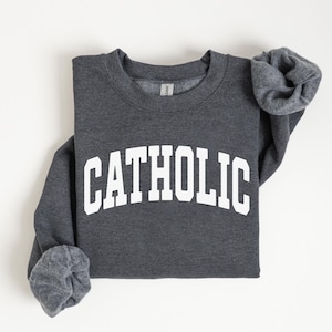 Peut inclure: Un sweat-shirt gris fonc&eacute; avec le mot "CATHOLIC" imprim&eacute; en grandes lettres blanches sur la poitrine. Le sweat-shirt est pli&eacute;, avec les manches partiellement visibles. Le fond est blanc uni.