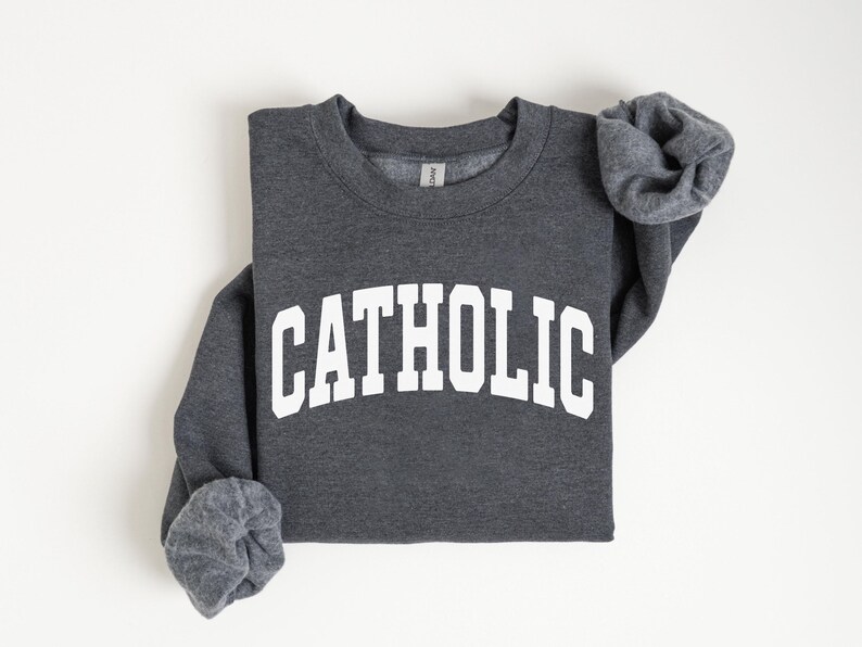Peut inclure: Un sweat-shirt gris fonc&eacute; avec le mot "CATHOLIC" imprim&eacute; en grandes lettres blanches sur la poitrine. Le sweat-shirt est pli&eacute;, avec les manches partiellement visibles. Le fond est blanc uni.