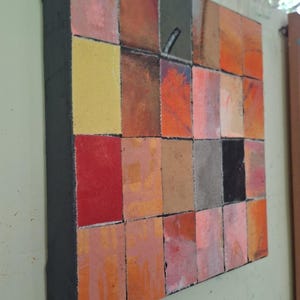 May include: A square canvas painting featuring a grid of rectangular blocks in various shades of orange, red, yellow, pink, gray, and black. The surface has a textured appearance with visible brushstrokes and a thick, dark-painted side edge.