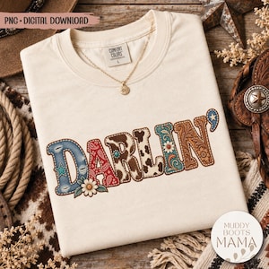 Könnte beinhalten: Ein cremefarbenes Kurzarm-T-Shirt mit dem Wort DARLIN' in großen Buchstaben. Jeder Buchstabe hat ein einzigartiges Muster, darunter Denim, rotes Paisley, Kuhflecken, türkisfarbene Blumen und braune Lederprägungen mit kleinen Blüten.