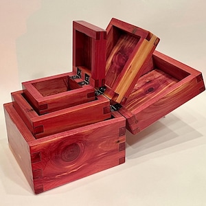 May include: A set of four wooden boxes, each with a hinged lid, crafted from a rich reddish-brown wood. The boxes are nested inside each other, creating a tiered effect. The wood grain is visible on the surface of the boxes.